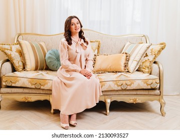 an attractive brunette woman in an elegant beige dress sits on a classic retro sofa. vintage clothes and interior. production of furniture to order.