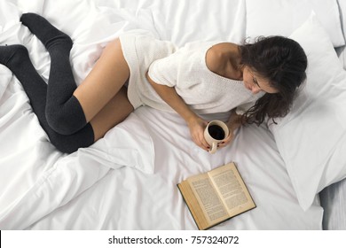 Attractive brunette woman reading book and holding cup of coffee (tea) in bed during the morning. Top view.