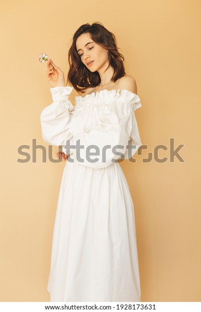 Attractive brunette woman wear white dress with\
elastic bands on shoulder while standing on beige background.\
Street fashion. Model wear sexy outfit and hold flower. Details of\
everyday elegant\
look.