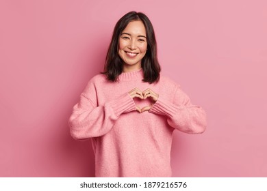 Attractive brunette young Asian woman feels happy and romantic shapes heart gesture expresses tender feelings wears casual jumper poses against pink background. People affection and care concept - Shutterstock ID 1879216576