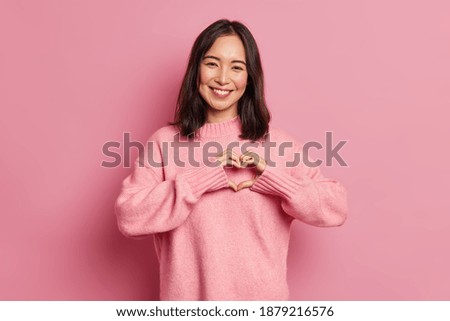 Attractive brunette young Asian woman feels happy and romantic shapes heart gesture expresses tender feelings wears casual jumper poses against pink background. People affection and care concept