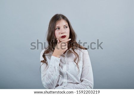 Attractive brunette young woman in fashion white shirt keeping hand on chin, thinking about something isolated on gray background in studio. People sincere emotions, lifestyle concept.