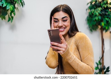 Attractive Brunette Young Woman, Holding Hand To Face, Scrolling Through Her Mobile Phone At Home.