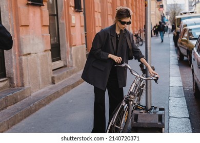 Attractive Business Woman Come To Sits On Bike. Girl Wear Grey Jacket Or Blazer, Black Pants, Eyeglasses. Modern Business Woman Ride To Work On Vehicle. Way To Office. Riding Bicycle To Work.