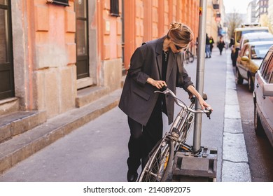 Attractive Business Woman Come To Sits On Bike. Girl Wear Grey Jacket Or Blazer, Black Pants, Eyeglasses. Modern Business Woman Ride To Work On Vehicle. Riding Bicycle To Work. Focus On Bike.