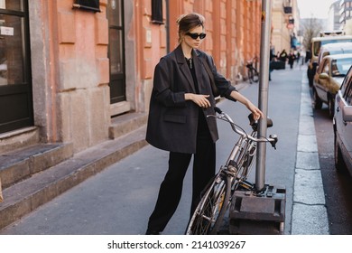 Attractive Business Woman Come To Sits On Bike. Girl Wear Grey Jacket Or Blazer, Black Pants, Eyeglasses. Modern Business Woman Ride To Work On Vehicle. Riding Bicycle To Work. Focus On Bike.