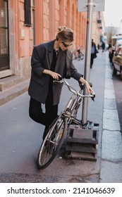 Attractive Business Woman Come To Sits On Bike. Girl Wear Grey Jacket Or Blazer, Black Pants, Eyeglasses. Modern Business Woman Ride To Work On Vehicle. Riding Bicycle To Work. Focus On Bike.