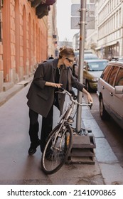 Attractive Business Woman Come To Sits On Bike. Girl Wear Grey Jacket Or Blazer, Black Pants, Eyeglasses. Modern Business Woman Ride To Work On Vehicle. Riding Bicycle To Work. Focus On Bike.