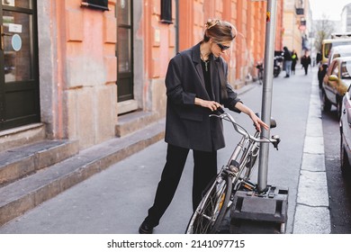 Attractive Business Woman Come To Sits On Bike. Girl Wear Grey Jacket Or Blazer, Black Pants, Eyeglasses. Modern Business Woman Ride To Work On Vehicle. Way To Office. Riding Bicycle To Work.