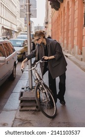 Attractive Business Woman Come To Sits On Bike. Girl Wear Grey Jacket Or Blazer, Black Pants, Eyeglasses. Modern Business Woman Ride To Work On Vehicle. Riding Bicycle To Work. Focus On Bike.