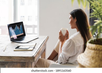 Attraktive Geschäftsfrau sitzt am Tisch vor dem Laptop und verwendet Smartphone für die Arbeit. Schönes Brunette Mädchen analysiert den Aktienmarkt während zu Hause. Millenniums-Frauen im Internet.