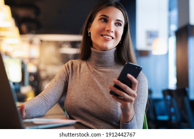 Attractive Business Woman Sits At Table In Front Of Laptop And Talks On Mobile Phone, Negotiates On The Phone. Beautiful Brunette Female Uses Gadgets For Work. Online Training, Remote Work From Home