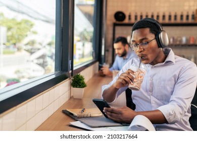 Attractive Businessman Listening Music With Headphones Sitting Near Windows At Coffee Shop Or Cafe. Holding A Plastic Cup Of Ice Coffee. Chill Out Life And Digital Lifestyle. Drinking And Relaxation