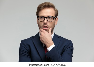 attractive businessman wearing suit and eyeglasses standing and looking away pensive and shocked on gray studio background
