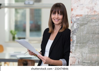Attractive businesswoman dealing with paperwork leaning against a wall in the office holding a document in her hands smiling at the camera