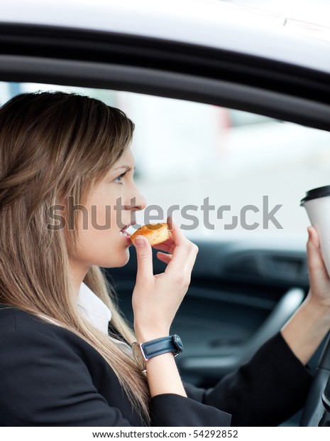 Attractive businesswoman eating and holding a\
drinking cup while driving to\
work