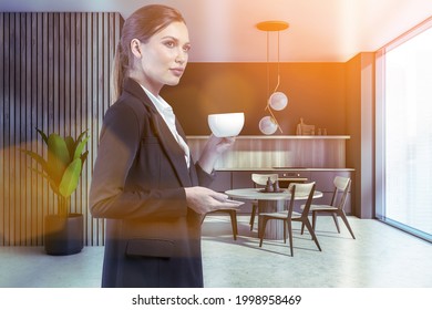 Attractive Businesswoman In Formal Suit Is Drinking Coffee At Corporate Office Kitchen. Modern Kitchen Set And Dining Area As Facilities For Employees. Sun Rays Filling The Room. Panoramic Window