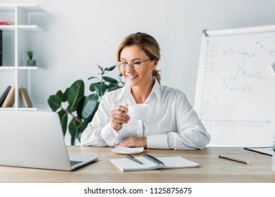 attractive businesswoman holding cup of coffee and looking at laptop in office