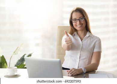 Attractive businesswoman looking at camera, showing thumbs up at workplace, pretty happy manager raising finger sitting at office desk with laptop, likes her job, acceptance approval sign, good idea