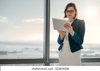 Attractive businesswoman using a digital tablet while standing in front of windows in an office building overlooking the city - Powered by Shutterstock