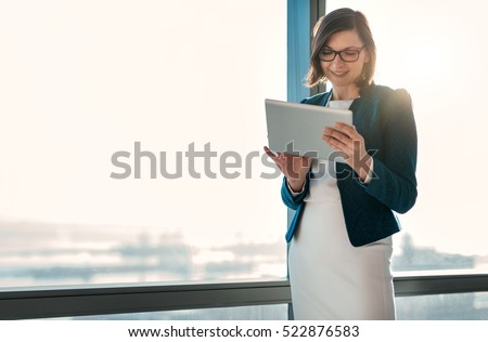 Attractive businesswoman using a digital tablet while standing in front of windows in an office building overlooking the city