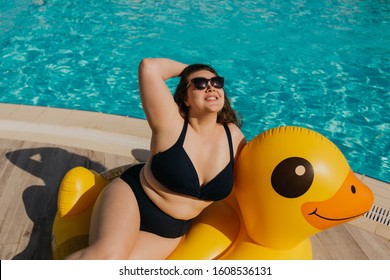 Attractive busty curvy woman in sunglasses and black swimsuit resting by the pool with yellow inflatable duck. Stylish accessories, fashion for plus size. Body positive, natural real beauty, resort