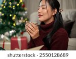 An attractive, calm Asian woman in a cozy red sweater enjoys a hot drink, cocoa or coffee, gently smelling it with her eyes closed, spending the Christmas holiday peacefully at home alone.