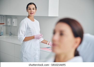 Attractive Calm Beautician In A Lab Coat And Latex Gloves Standing Behind A Dark-haired Patient