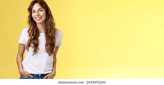 Attractive carefree confident friendly-looking charming feminine ubran woman long curly hair smiling toothy pleased tilt head intrigued hold hands pockets self-assured pose stand yellow background