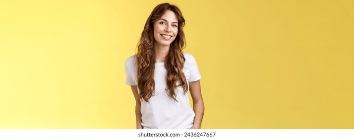 Attractive carefree confident friendly-looking charming feminine ubran woman long curly hair smiling toothy pleased tilt head intrigued hold hands pockets self-assured pose stand yellow background