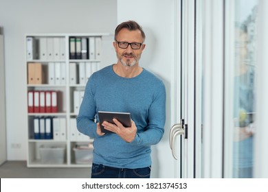 Attractive casual businessman using a tablet in the office as he stands alongside a glass entrance door looking at the camera