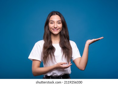 Attractive caucasian or arab brunette girl in a white t-shirt showing a copy space on her palm for advertising isolated on a blue studio background. - Shutterstock ID 2119256330