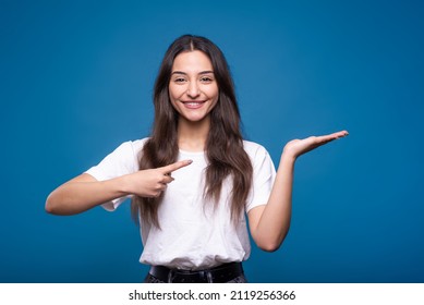 Attractive caucasian or arab brunette girl in a white t-shirt pointing her finger at the copy space on the palm for advertising isolated on a blue studio background. - Shutterstock ID 2119256366