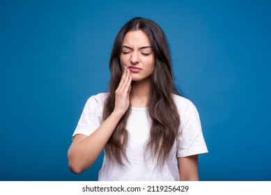 Attractive caucasian or arab brunette girl in a white t-shirt touching her cheek because of a toothache isolated on a blue studio background.