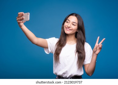 Attractive Caucasian Or Arab Brunette Girl In A White T-shirt Taking A Selfie On A Mobile Phone Camera And Showing A Peace Gesture Isolated On A Blue Studio Background.