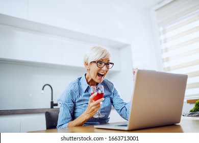 Attractive Caucasian Blond Senior Woman Sitting At Dining Table, Drinking Wine, Looking At Laptop And Cheering For Achievement.