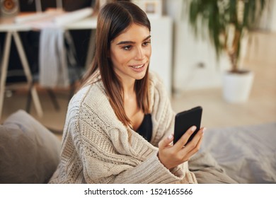 Attractive caucasian brunette dressed in beige sweater sitting on bed in bedroom and using smart phone. - Shutterstock ID 1524165566