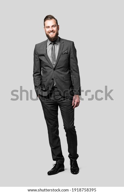 Attractive caucasian businessman\
with a beard\
and a friendly smile wearing a grey suit and tie, standing casually\
with one hand in his\
pocket.