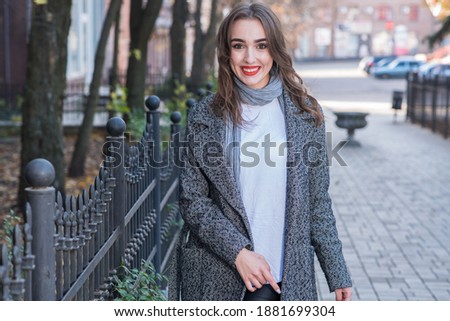 Attractive Caucasian elegant young brunette woman with red lips, wearing a coat, posing, smiling and looking at the camera while walking outdoors in autumn. Style, fashion, elegance.