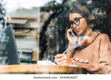 Attractive caucasian female digital nomad freelancer talking on cell phone and working on laptop making notes in a notebook while sitting in modern coffee shop. Woman working remote on cafe restaurant