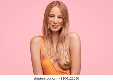 Attractive Caucasian female has festive make up, decorated with sparkles on face and hair, poses for fashion magazine, shows her beauty, stands alone against pink background. Glitter on body