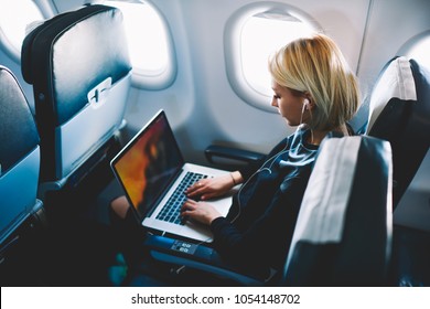Attractive caucasian female passenger of airplane sitting in comfortable seat listening music in earphones while working at modern laptop computer with mock up area using wireless connection on board