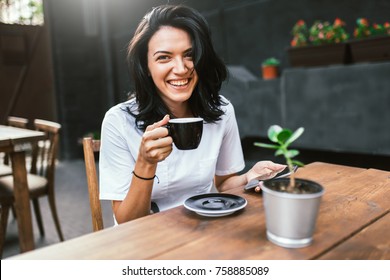 Attractive Caucasian female with pleasant cheerful smile sitting at terrace cafe, drinking coffee and enjoying online communication using free wireless internet connection on her smart phone. 