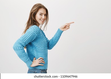 attractive caucasian female standing in profile while pointing right with index finger and looking at camera with bright smile, over gray background. Girl dare friend flirt with woman who sits alone