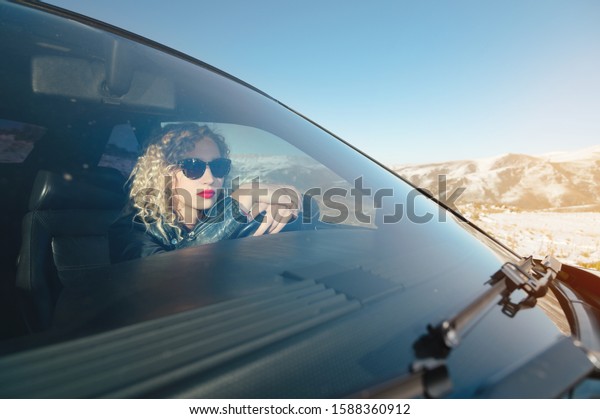Attractive Caucasian girl in sunglasses and a\
leather jacket sits at the wheel of a sports retro car with a\
leather interior. Woman driving\
concept