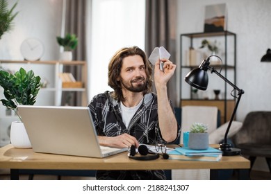 Attractive Caucasian Man In Casual Outfit Sitting At Desk With Modern Laptop And Playing With Paper Plane. Male Boring At Work. Unproductive Concept.