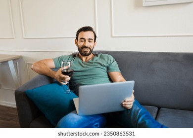 Attractive caucasian man drinking wine during a video call and having a fun virtual date with his girlfriend at home - Powered by Shutterstock