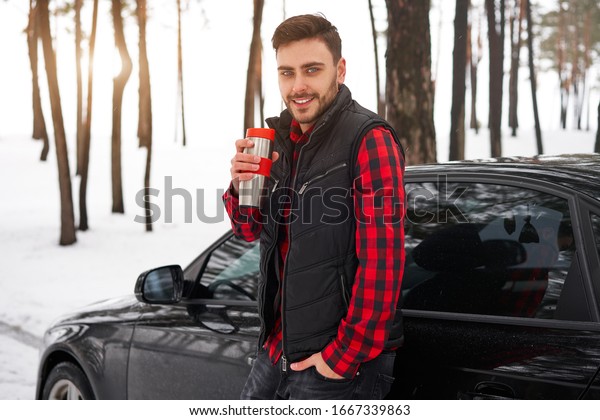 Attractive Caucasian man stands winter forest near\
car drink hot beverage thermos. Winter holiday road trip. Cold\
season. Portrait young handsome unshaved driver dressed red\
checkered shirt vest