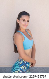 Attractive Caucasian millennial woman wearing sportswear in blue tones and posing after a fitness session looking at the camera and smiling, athletic person showing off her fit and beautiful physique