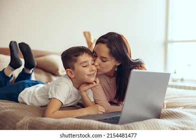 Attractive Caucasian Mother Kissing Small Teen Son Lying On Bed, Looking At Laptop Screen.